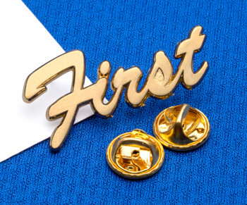 Gold script lapel pin reading First with two butterfly clutch backings on a blue textured background.