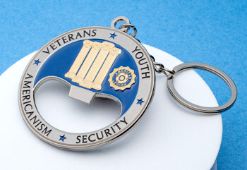 Circular silver and blue enamel bottle opener keychain featuring the American Legion emblem, four pillars, and text: Veterans, Youth, Americanism, Security.