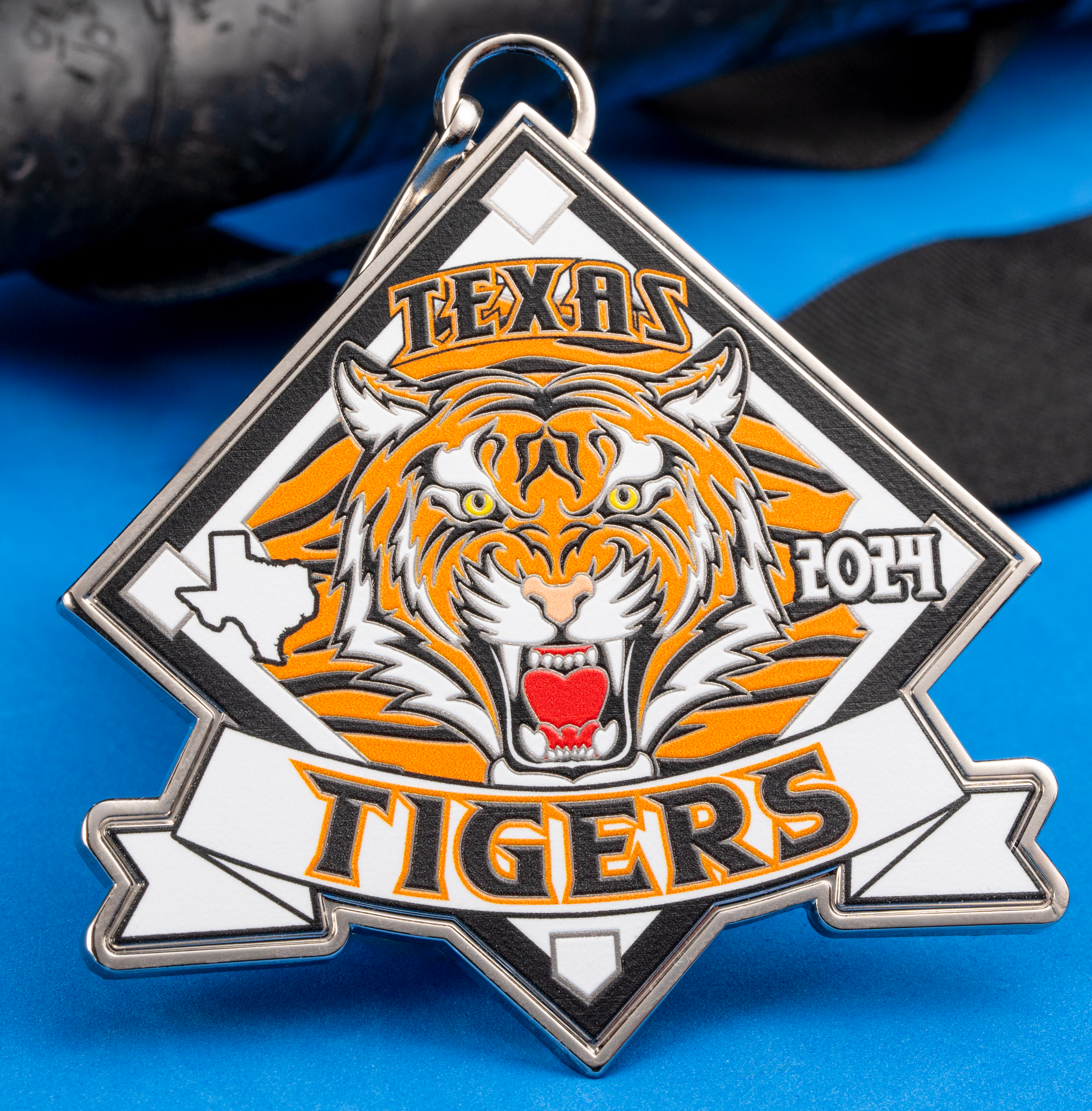 Diamond-shaped custom medal featuring a snarling tiger head, the state of Texas, and 'Texas Tigers 2024' text on a blue surface with a baseball bat.