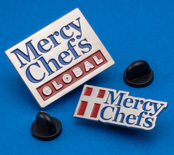 Two Mercy Chefs branded enamel lapel pins with blue lettering and red accents on a blue background with black rubber clutch backings.