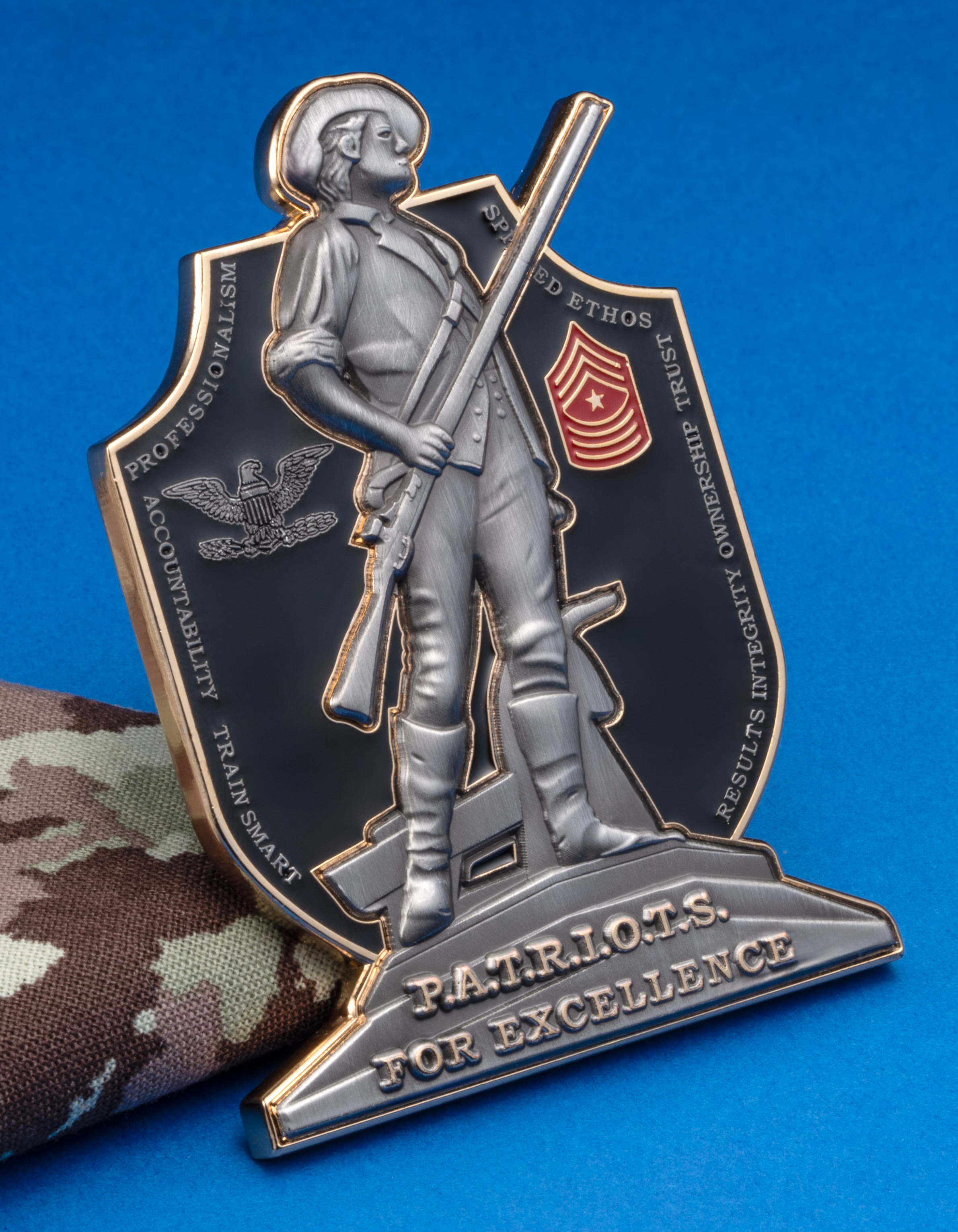Shield-shaped metal challenge coin featuring a 3D relief of a Minuteman holding a rifle, an eagle emblem, and a chevron rank insignia against a black background