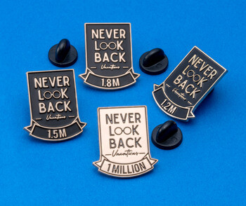 Four enamel lapel pins with 'Never Look Back Vacations' text and various milestone markers including 1 Million, 1.2M, 1.5M, and 1.8M on a blue background.