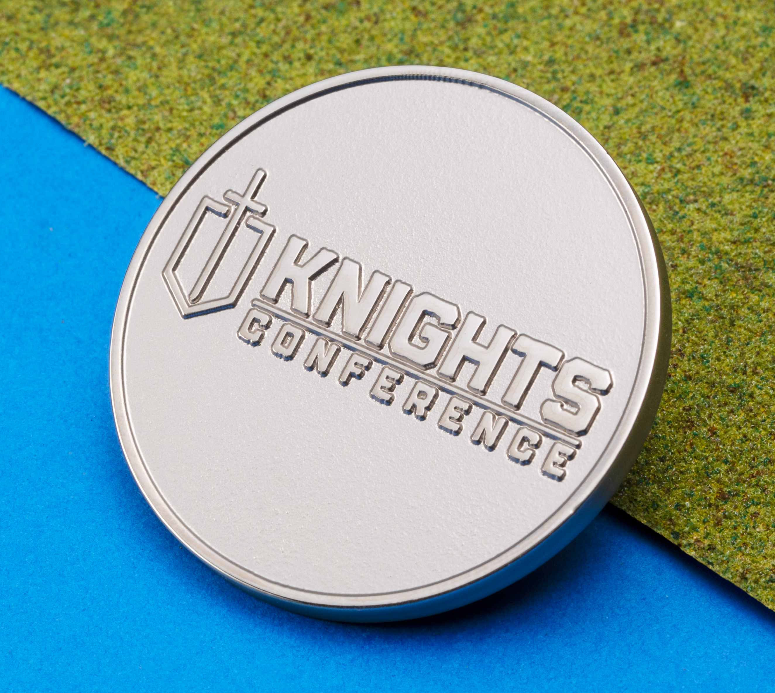 A round silver metal coin featuring the embossed Knights Conference logo and a shield icon, resting on a blue and green textured background.
