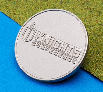 A round silver metal coin featuring the embossed Knights Conference logo and a shield icon, resting on a blue and green textured background.