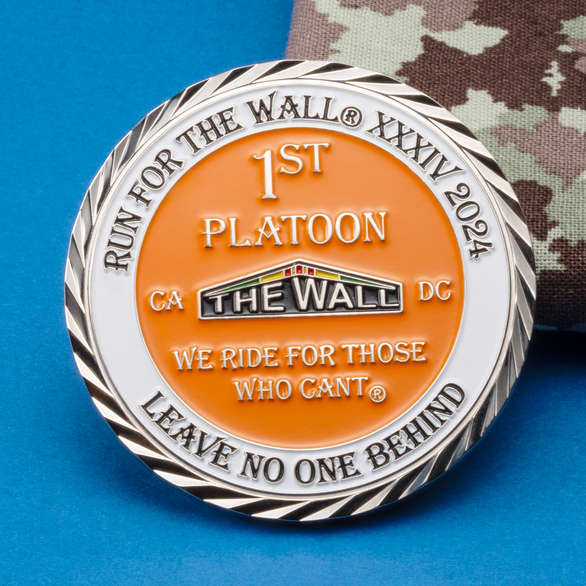 Enamel challenge coin for Run For The Wall XXXIV 2024, 1st Platoon, with orange and white enamel on a blue background next to camouflage fabric.