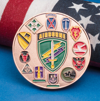 A circular rose gold challenge coin featuring various U.S. Army unit insignia and a central Airborne shield, resting against a rolled American flag.