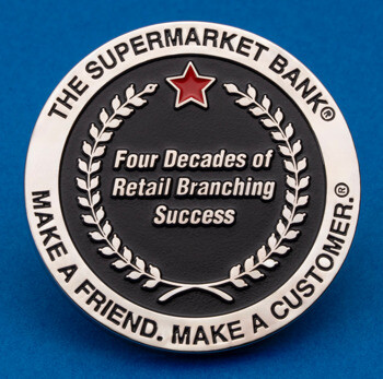 Circular silver lapel pin for The Supermarket Bank featuring a red star, laurel wreath, and black center with anniversary text on a blue background.