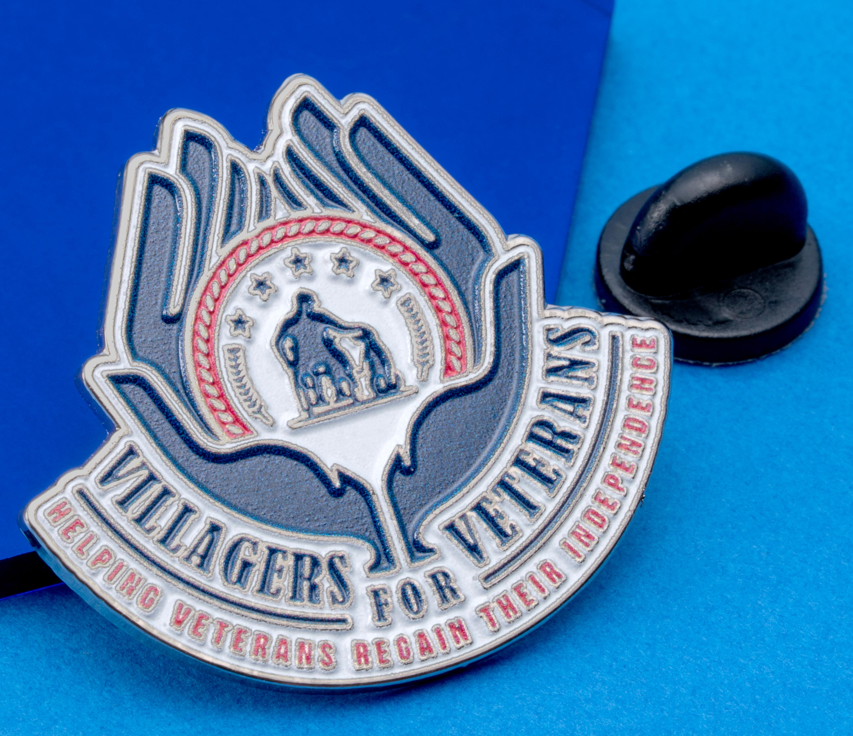 Soft enamel lapel pin for Villagers For Veterans featuring two hands holding a central emblem with stars and a figure, displayed on a blue background.