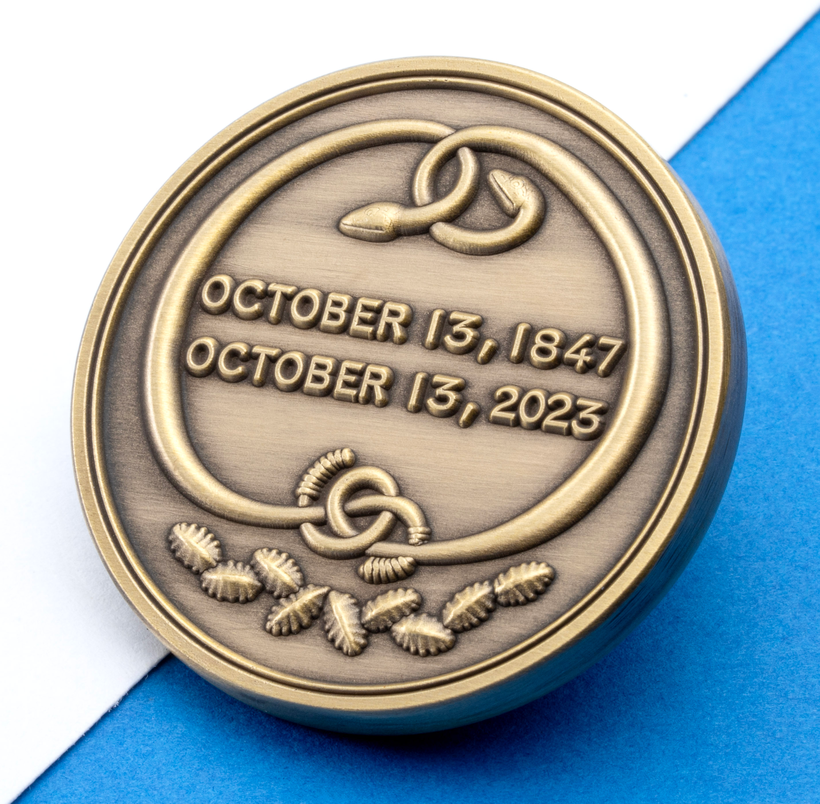 Antiqued bronze circular medallion featuring the dates October 13, 1847 and October 13, 2023 with interlaced snake and knot symbols.