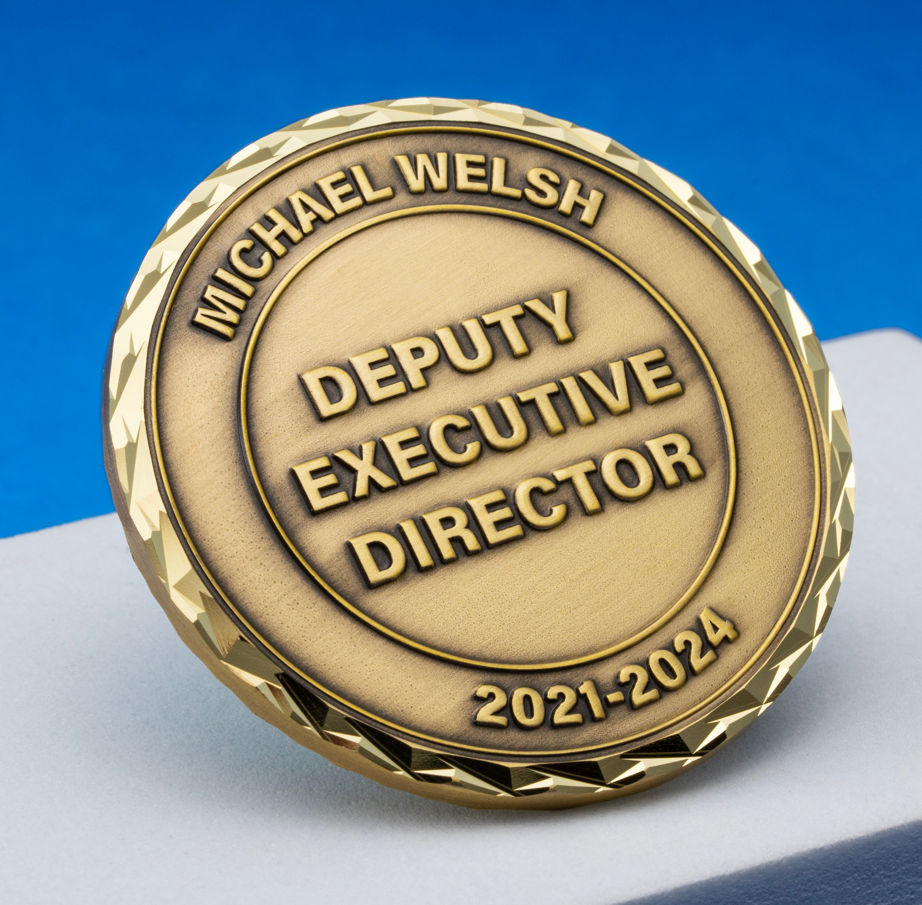 Antiqued gold coin inscribed with Michael Welsh Deputy Executive Director 2021-2024 featuring a diamond-cut edge on a white display block.