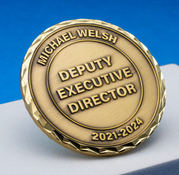 Antiqued gold coin inscribed with Michael Welsh Deputy Executive Director 2021-2024 featuring a diamond-cut edge on a white display block.