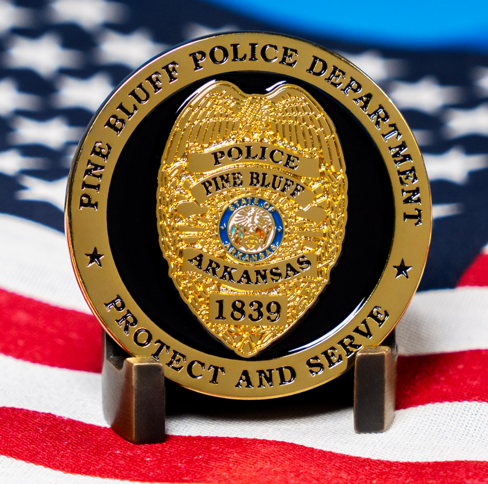A gold and black Pine Bluff Police Department challenge coin featuring a police badge emblem, resting on a folded American flag.