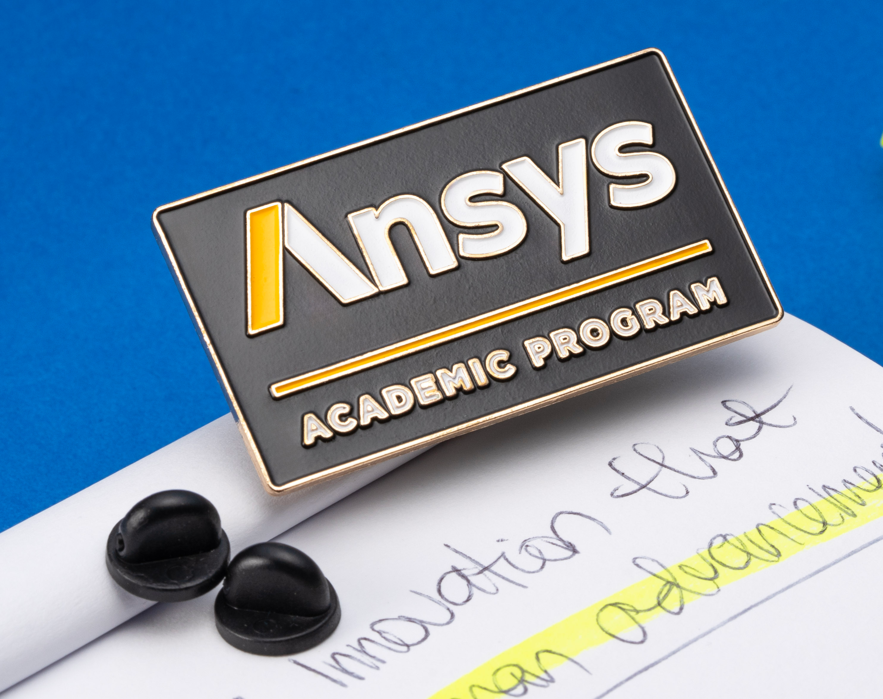 A rectangular black lapel pin with the Ansys logo and Academic Program text resting on a notepad next to a yellow highlighter.
