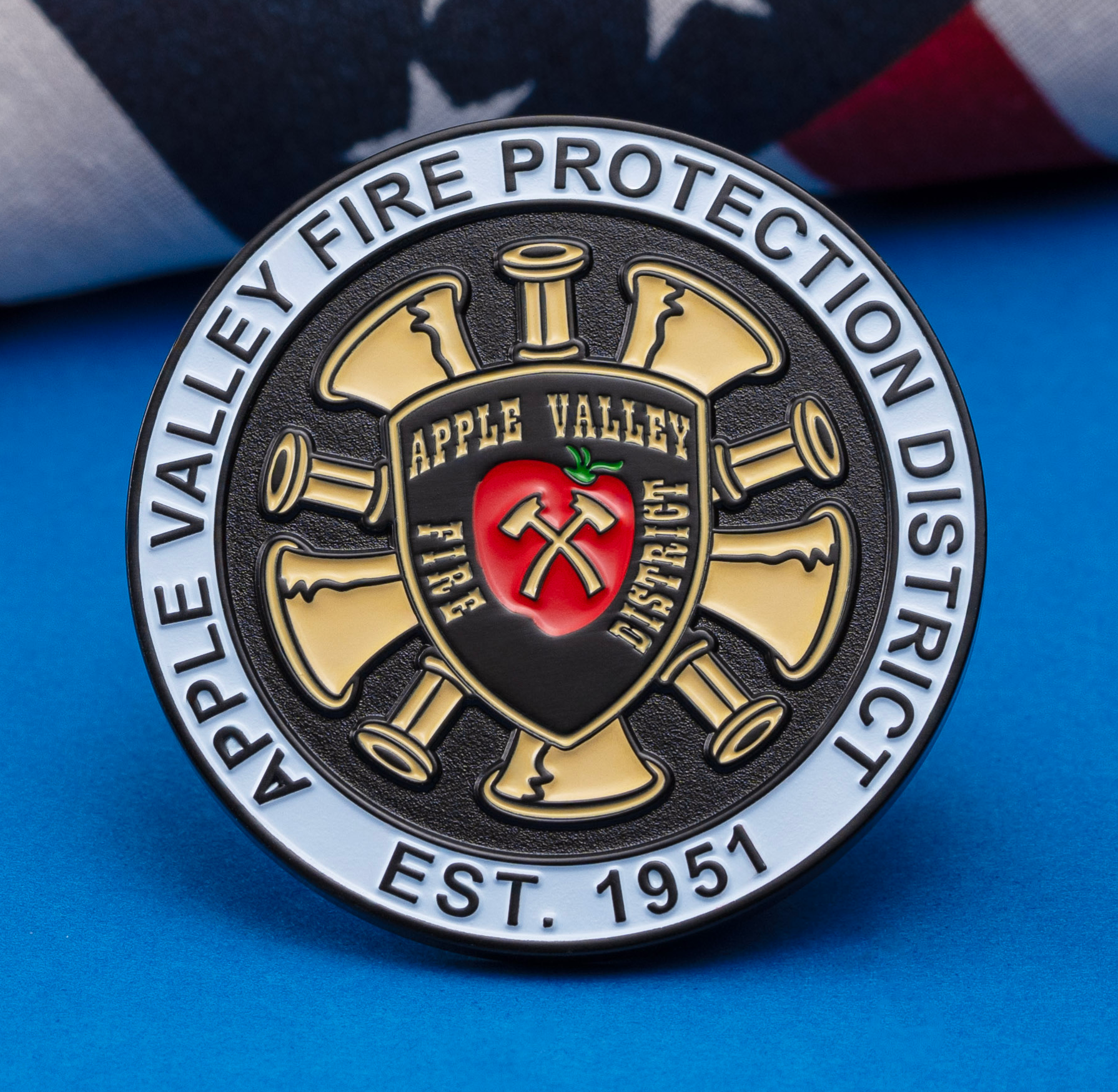 A circular challenge coin for Apple Valley Fire Protection District featuring a red apple and firefighting axes, set against a blue background with a US flag.