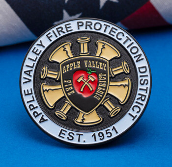 A circular challenge coin for Apple Valley Fire Protection District featuring a red apple and firefighting axes, set against a blue background with a US flag.