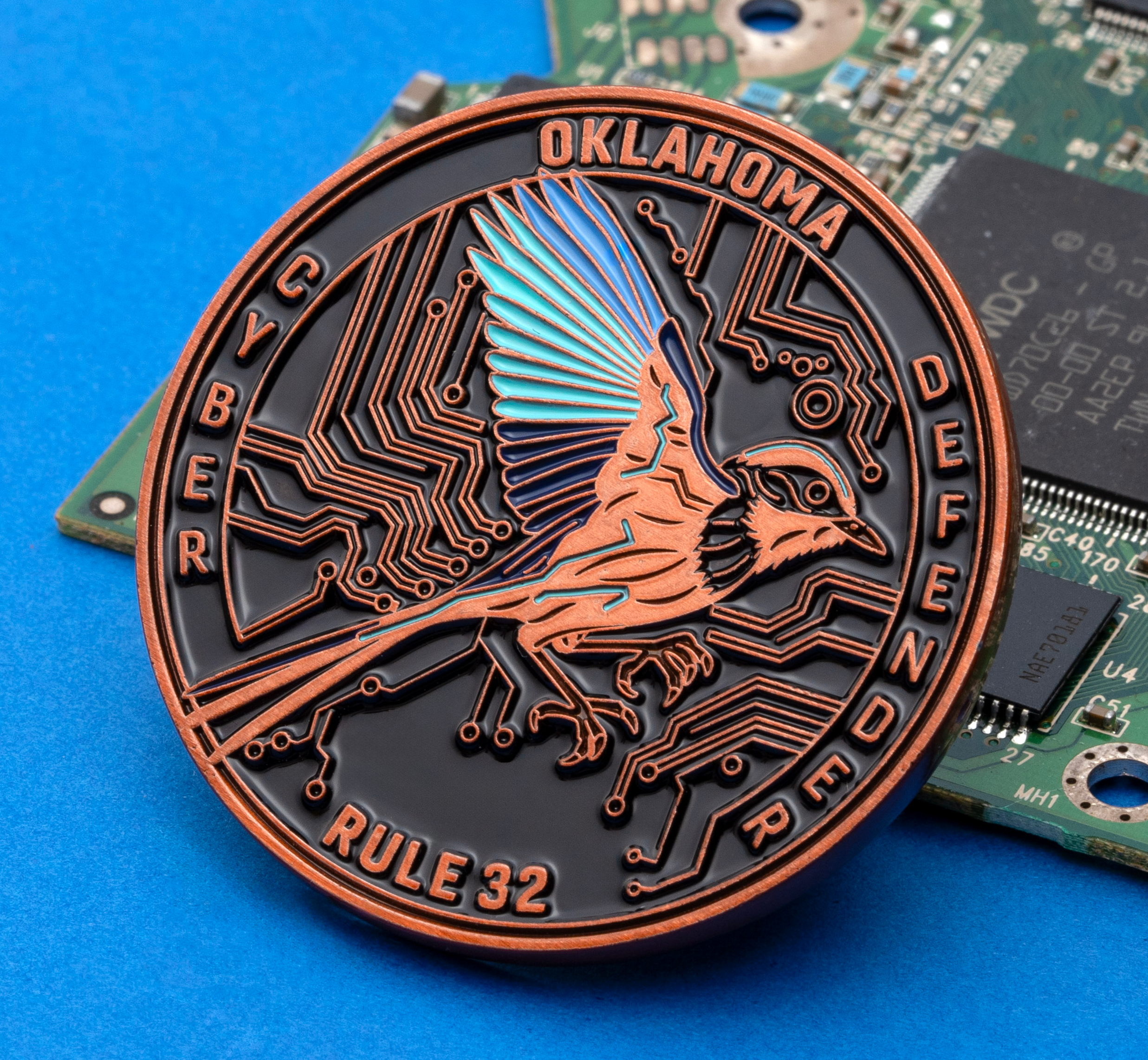 Circular copper and black challenge coin with a bird and circuit board design, labeled Oklahoma Cyber Defender Rule 32, resting on a green circuit board.