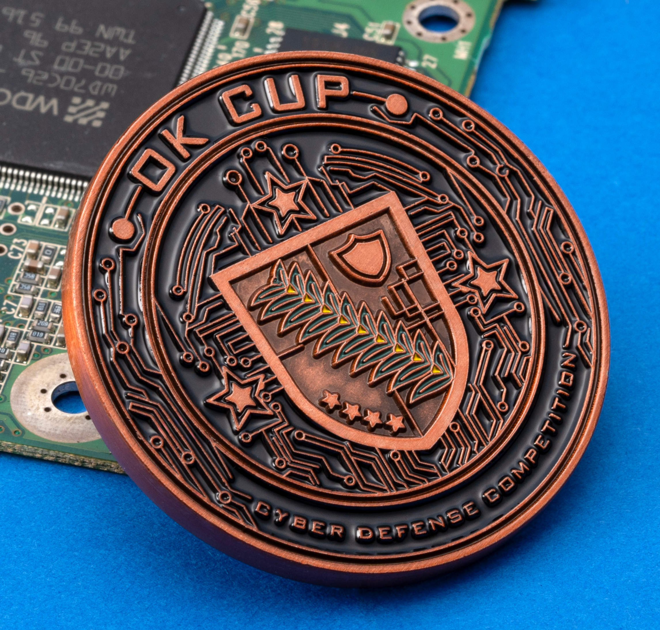 Antique copper OK Cup challenge coin featuring a shield and circuit board design, resting on a green motherboard against a blue background.