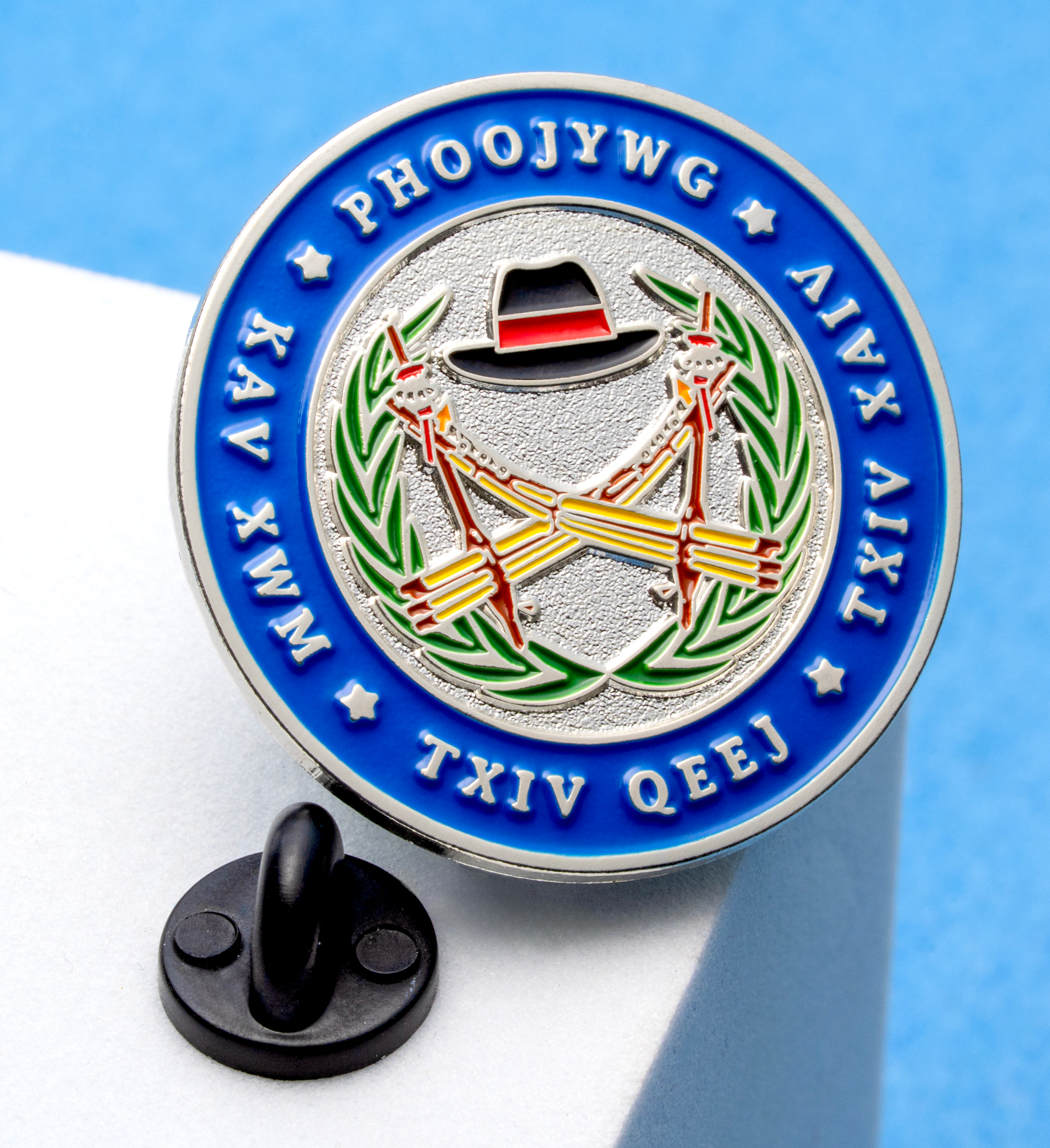 Circular soft enamel lapel pin with blue outer ring containing white text, featuring a center emblem with a black hat, crossed instruments, and green laurels.