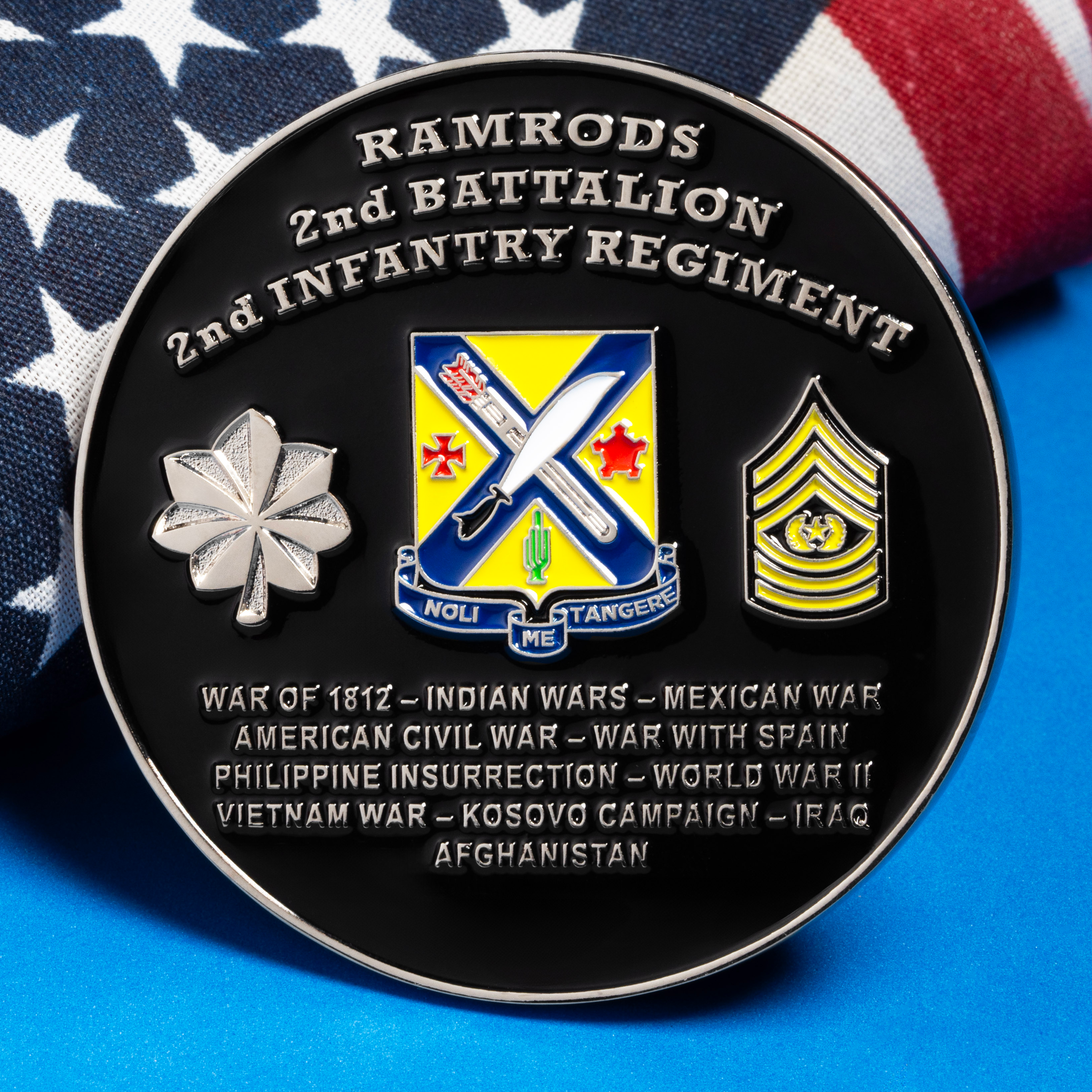 Black challenge coin for the 2nd Battalion 2nd Infantry Regiment resting against a rolled American flag on a blue surface.