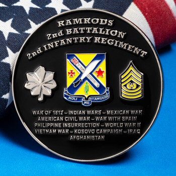 Black challenge coin for the 2nd Battalion 2nd Infantry Regiment resting against a rolled American flag on a blue surface.