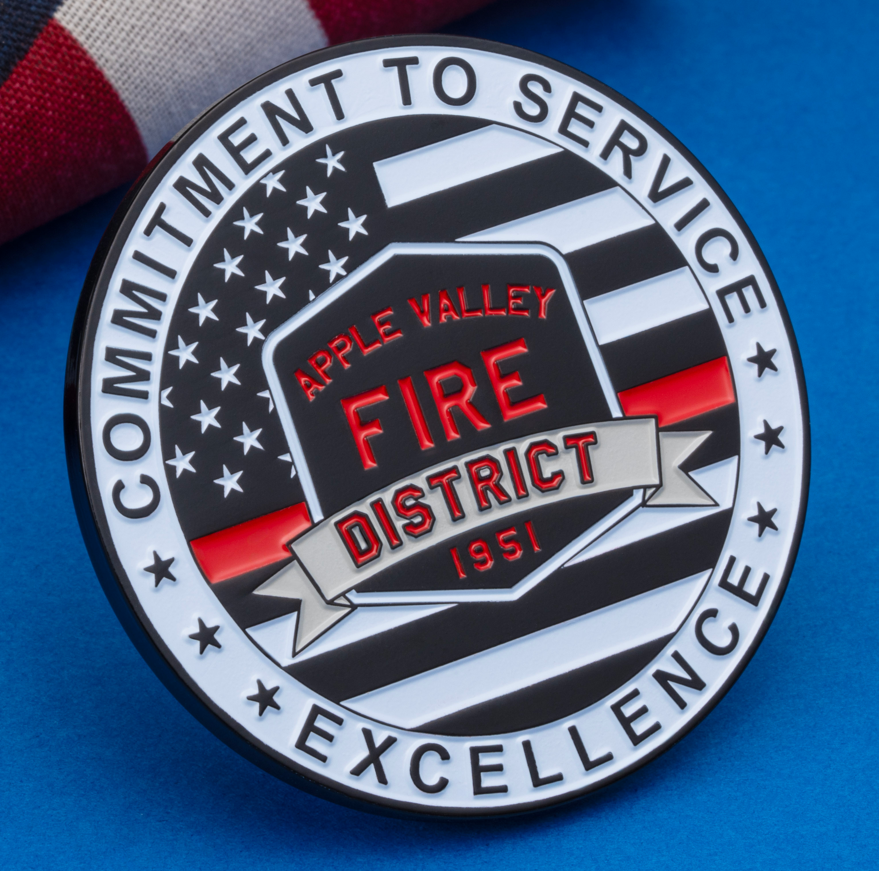 Circular Apple Valley Fire District challenge coin with American flag design and red line detail, placed next to a folded US flag on a blue surface.