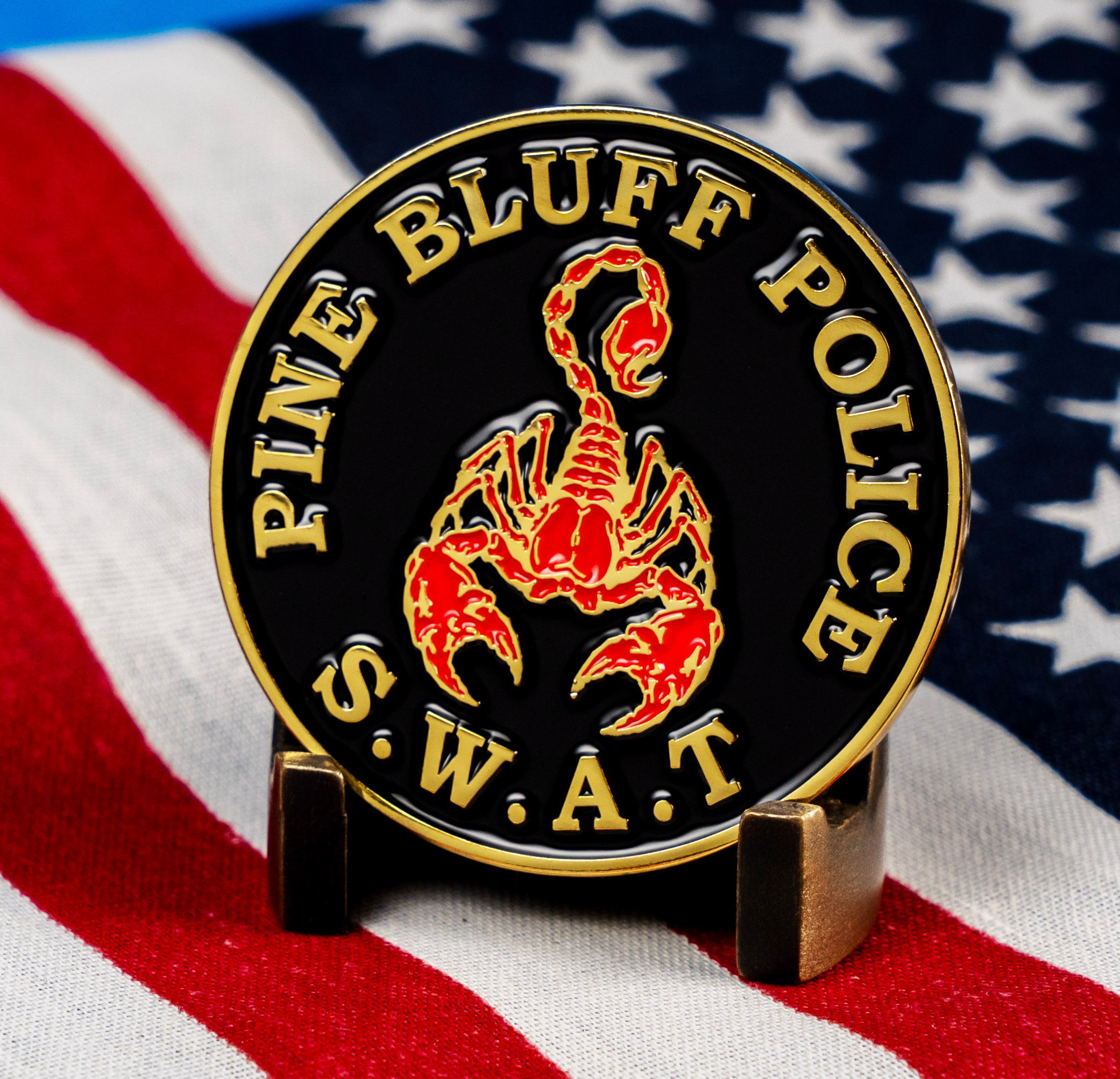 A circular black and gold challenge coin featuring a red and gold scorpion, with text Pine Bluff Police S.W.A.T., displayed on an American flag.