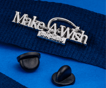Silver Make-A-Wish San Diego lapel pin attached to a navy blue fabric strap with two black rubber clutch backings on a blue surface.