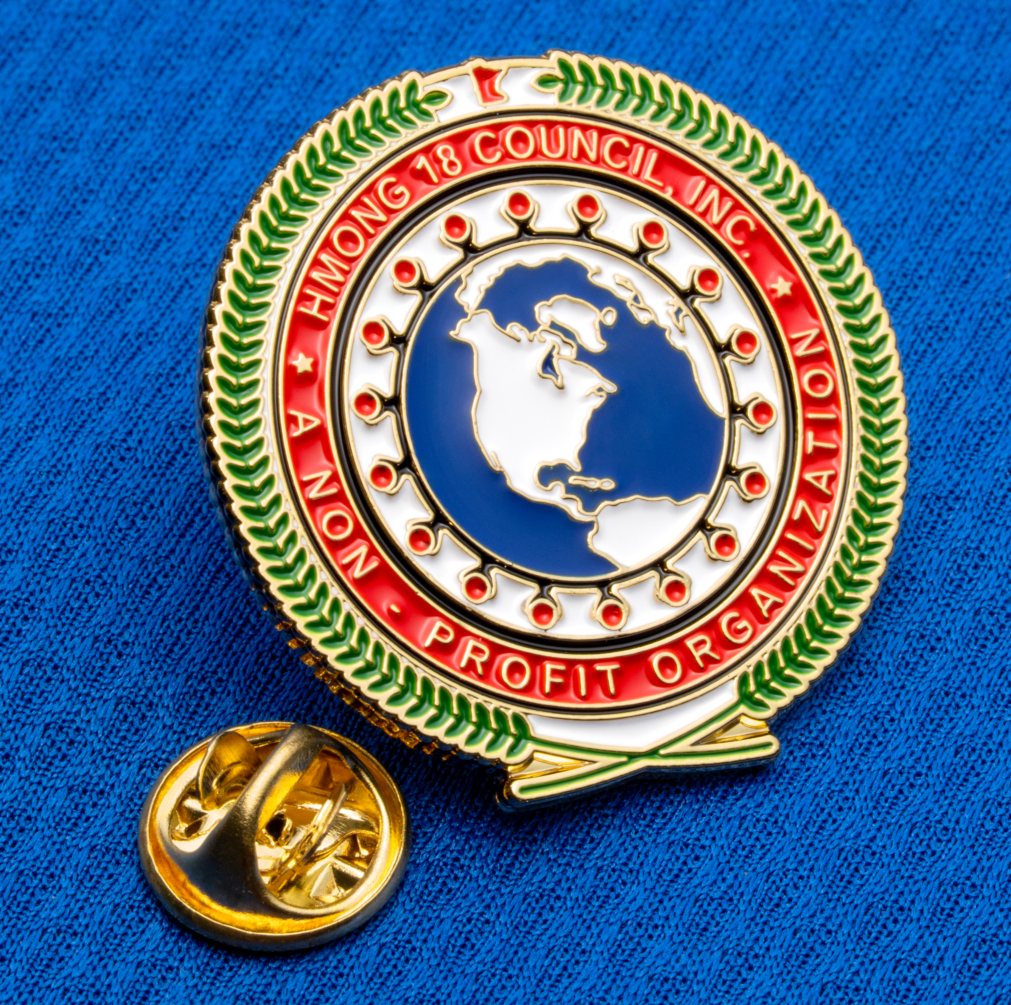 Enamel lapel pin for Hmong 18 Council Inc. featuring a globe, laurel wreath, and red border on a blue fabric background.