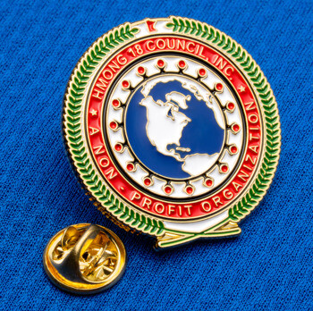 Enamel lapel pin for Hmong 18 Council Inc. featuring a globe, laurel wreath, and red border on a blue fabric background.