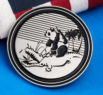 Circular silver and black challenge coin featuring a panda riding an alligator with a sunset and palm tree background, placed next to an American flag.