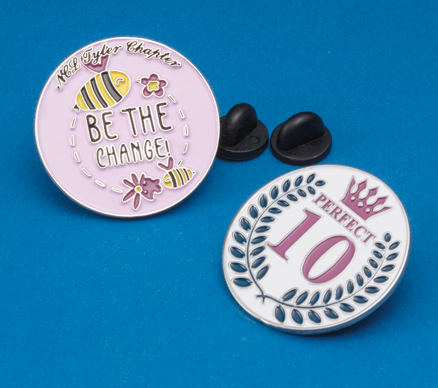 Enamel lapel pins featuring a bee design with 'Be the Change' and a laurel wreath design with 'Perfect 10'.