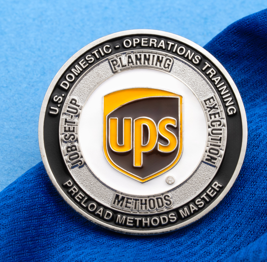 Circular challenge coin featuring the UPS logo, silver and black outer ring, and white inner circle with training department text.
