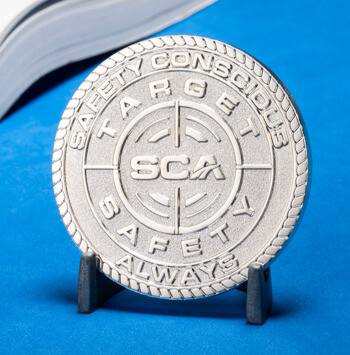 SCA Target Safety Always silver challenge coin with a crosshair emblem and rope border on a blue background next to a book.