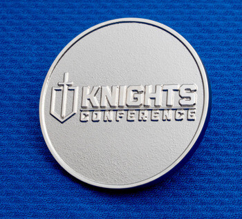 Silver Knights Conference challenge coin featuring a sword and shield emblem on a blue textured background.