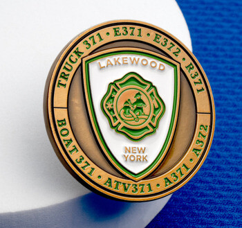 Circular brass and enamel Lakewood New York fire department challenge coin with a green Maltese cross emblem and unit numbers on a blue background.