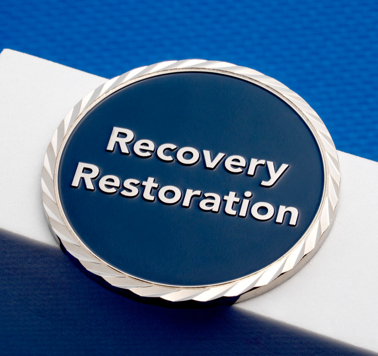 A silver challenge coin featuring a dark blue enamel center with 'Recovery Restoration' text and a textured rope-style border.