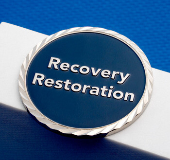 A silver challenge coin featuring a dark blue enamel center with 'Recovery Restoration' text and a textured rope-style border.