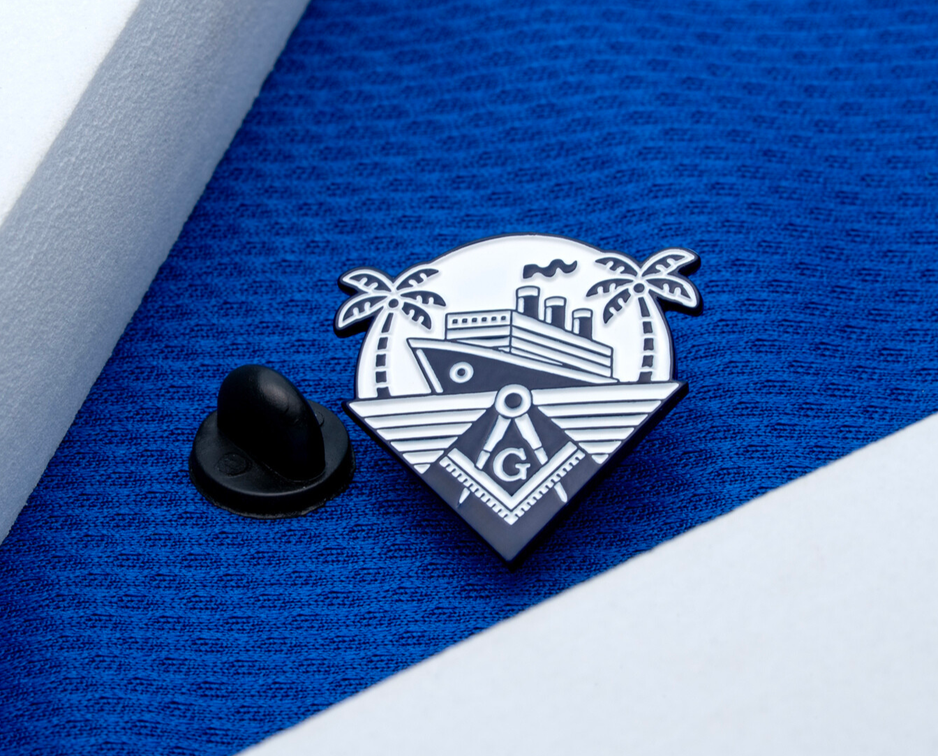 Enamel lapel pin featuring a cruise ship, palm trees, and a Masonic square and compass symbol on a blue fabric background.