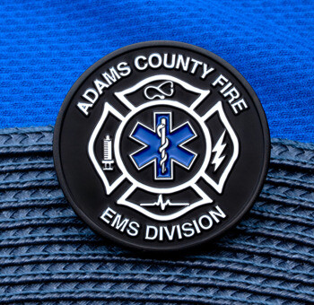 Circular Adams County Fire EMS Division challenge coin featuring a Star of Life and maltese cross on a black background.