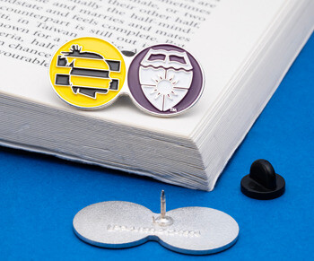Dual circle lapel pin featuring a yellow profile icon and a purple shield with sun design on an open book.
