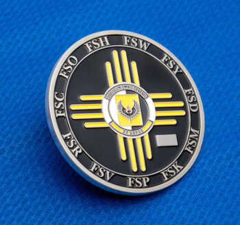Circular challenge coin with black and yellow Zia sun symbol and 27 SOFSS text on a blue fabric background.