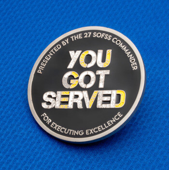 Black and silver challenge coin featuring the text You Got Served and 27 SOFSS Commander on a blue fabric background.