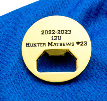 Gold circular bottle opener challenge coin engraved with 2022-2023 13U Hunter Mathews #23 on a blue fabric background.