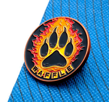 Circular lapel pin featuring a black animal paw print surrounded by orange and red flames with the text WAFFLE on a blue fabric background.