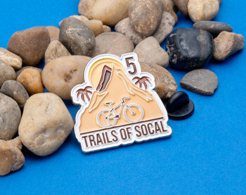 Soft enamel lapel pin featuring a mountain, bicycle, and palm trees with the text Trails of SoCal 5 resting on pebbles.