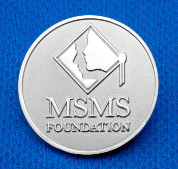 Silver MSMS Foundation challenge coin featuring a diamond emblem with two profile faces on a blue textured background.