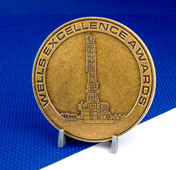 Antiqued gold Wells Excellence Awards challenge coin featuring an oil derrick design, displayed on a small stand against a blue and white background.