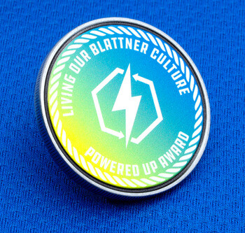 Circular challenge coin featuring a green and blue gradient with a white lightning bolt logo and 'Living Our Blattner Culture' text.