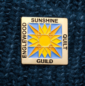 Englewood Sunshine Quilt Guild square lapel pin with a yellow and blue sunburst design pinned to a dark blue knit beanie.