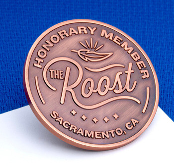 Antiqued copper lapel pin with 'The Roost' logo, reading 'Honorary Member Sacramento, CA' with a feather design, on a blue and white background.